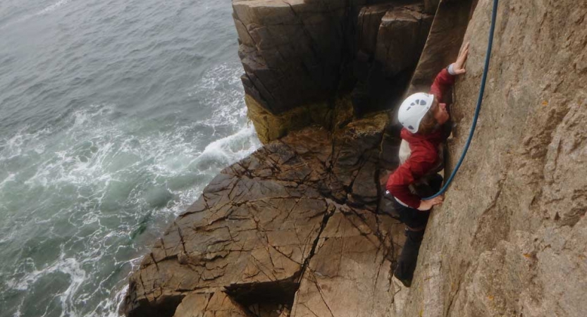 A person wearing safety gear is secured by ropes as they rock climb above the ocean.